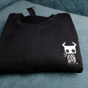 May include: Black sweatshirt with a small, pixelated white and gray character design on the front. The sweatshirt is folded neatly on a teal surface. The design appears to be a video game character.