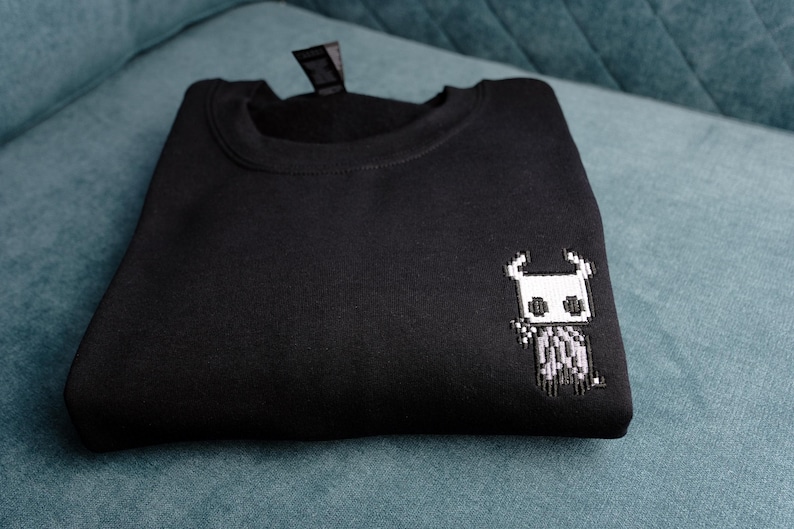 May include: Black sweatshirt with a small, pixelated white and gray character design on the front. The sweatshirt is folded neatly on a teal surface. The design appears to be a video game character.