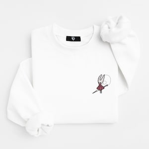 May include: White long-sleeve sweatshirt with a small embroidered design of a character in red and black. The character is holding a needle and a white circle. The sweatshirt has a small black tag on the collar.