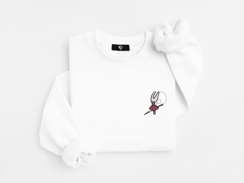 May include: White long-sleeve sweatshirt with a small embroidered design of a character in red and black. The character is holding a needle and a white circle. The sweatshirt has a small black tag on the collar.
