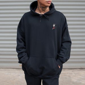 May include: Black hooded sweatshirt with a kangaroo pocket and a drawstring. The sweatshirt features a small embroidered design on the upper left chest. The model is wearing the sweatshirt with their hands in the pockets.