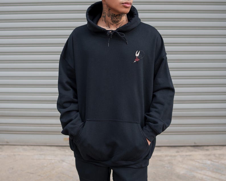 May include: Black hooded sweatshirt with a kangaroo pocket and a drawstring. The sweatshirt features a small embroidered design on the upper left chest. The model is wearing the sweatshirt with their hands in the pockets.