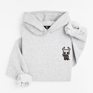 May include: A light gray hoodie with a pixelated character design on the front. The hoodie has a hood and long sleeves with rolled cuffs. The design is a black and white character.