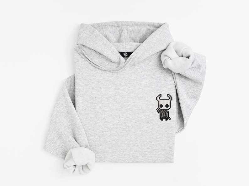 May include: A light gray hoodie with a pixelated character design on the front. The hoodie has a hood and long sleeves with rolled cuffs. The design is a black and white character.