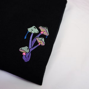 Peut inclure: T-shirt noir avec un motif brod&eacute; de champignons violets aux chapeaux verts.