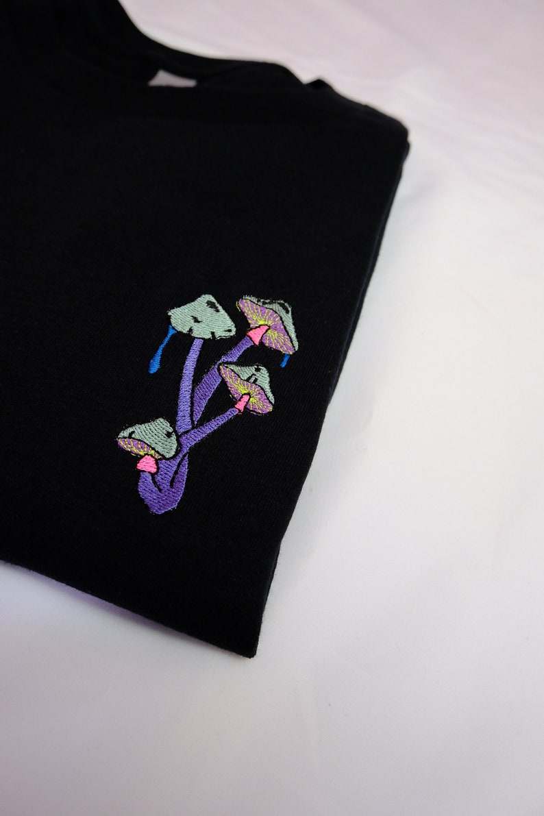 Peut inclure: T-shirt noir avec un motif brod&eacute; de champignons violets aux chapeaux verts.