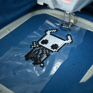 May include: An embroidery machine stitching a pixelated character design onto blue fabric. The design features a white and black figure with horns and a cape. The machine's needle is positioned above the fabric, ready to continue the embroidery process.