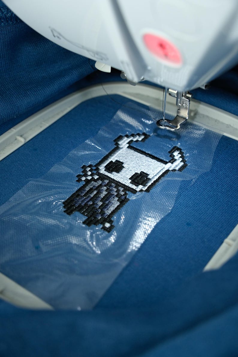 May include: An embroidery machine stitching a pixelated character design onto blue fabric. The design features a white and black figure with horns and a cape. The machine's needle is positioned above the fabric, ready to continue the embroidery process.