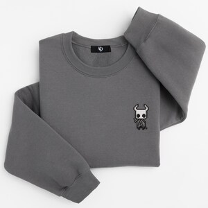 May include: A charcoal gray sweatshirt with a crew neck and long sleeves. A small pixelated character is embroidered on the front. The sweatshirt is folded neatly on a white surface.