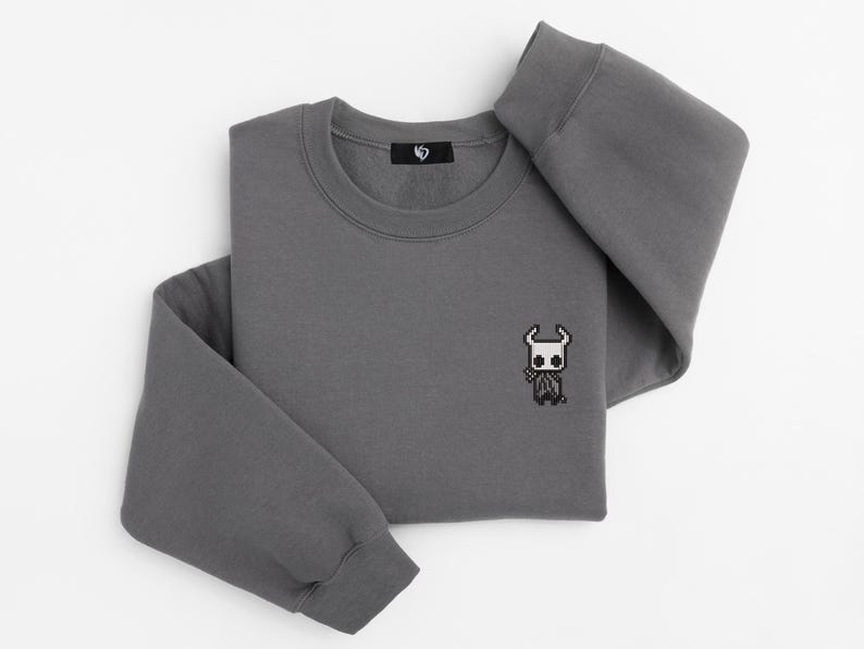 May include: A charcoal gray sweatshirt with a crew neck and long sleeves. A small pixelated character is embroidered on the front. The sweatshirt is folded neatly on a white surface.