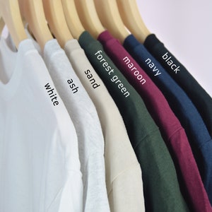 Puede incluir: Una fila de camisetas de varios colores, incluyendo blanco, ceniza, arena, verde bosque, granate, azul marino y negro. Las camisetas est&aacute;n colgadas en perchas de madera.