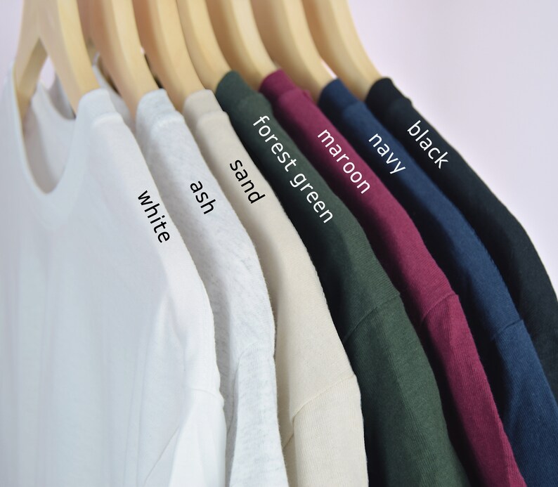 Puede incluir: Una fila de camisetas de varios colores, incluyendo blanco, ceniza, arena, verde bosque, granate, azul marino y negro. Las camisetas est&aacute;n colgadas en perchas de madera.