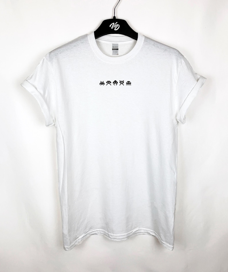 May include: White t-shirt with rolled-up sleeves, featuring a small black pixelated alien design across the chest. The shirt is hanging on a black hanger. The design is reminiscent of classic arcade games.
