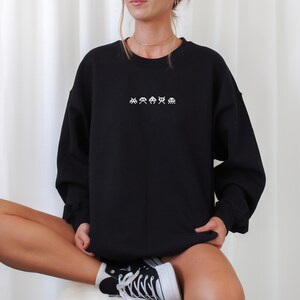 May include: Black crewneck sweatshirt with white pixelated alien graphics across the chest. The sweatshirt is long-sleeved and has a relaxed fit. The person is wearing black and white sneakers.