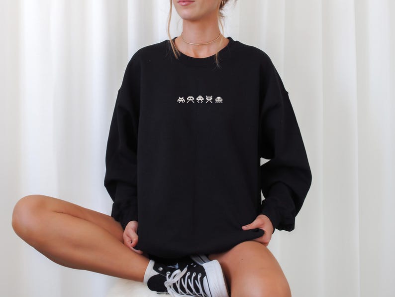 May include: Black crewneck sweatshirt with white pixelated alien graphics across the chest. The sweatshirt is long-sleeved and has a relaxed fit. The person is wearing black and white sneakers.