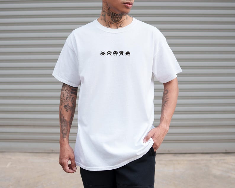 May include: White t-shirt with a crew neck featuring a small, black pixelated alien design across the chest. The shirt is a solid white color and appears to be made of a soft material. The model is wearing black shorts.