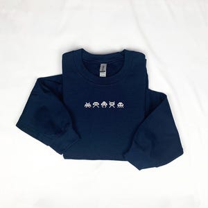 May include: A navy blue long-sleeved sweatshirt featuring a pixelated arcade game design in white. The crew neck sweatshirt is made of a soft material and is laid flat on a white surface.