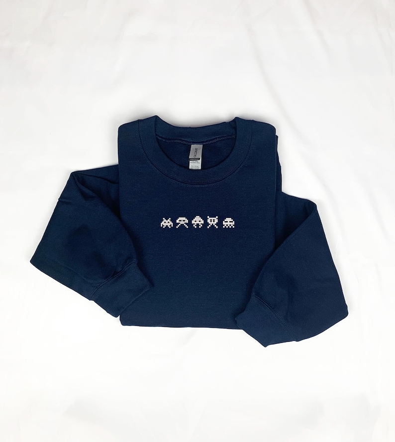 May include: A navy blue long-sleeved sweatshirt featuring a pixelated arcade game design in white. The crew neck sweatshirt is made of a soft material and is laid flat on a white surface.