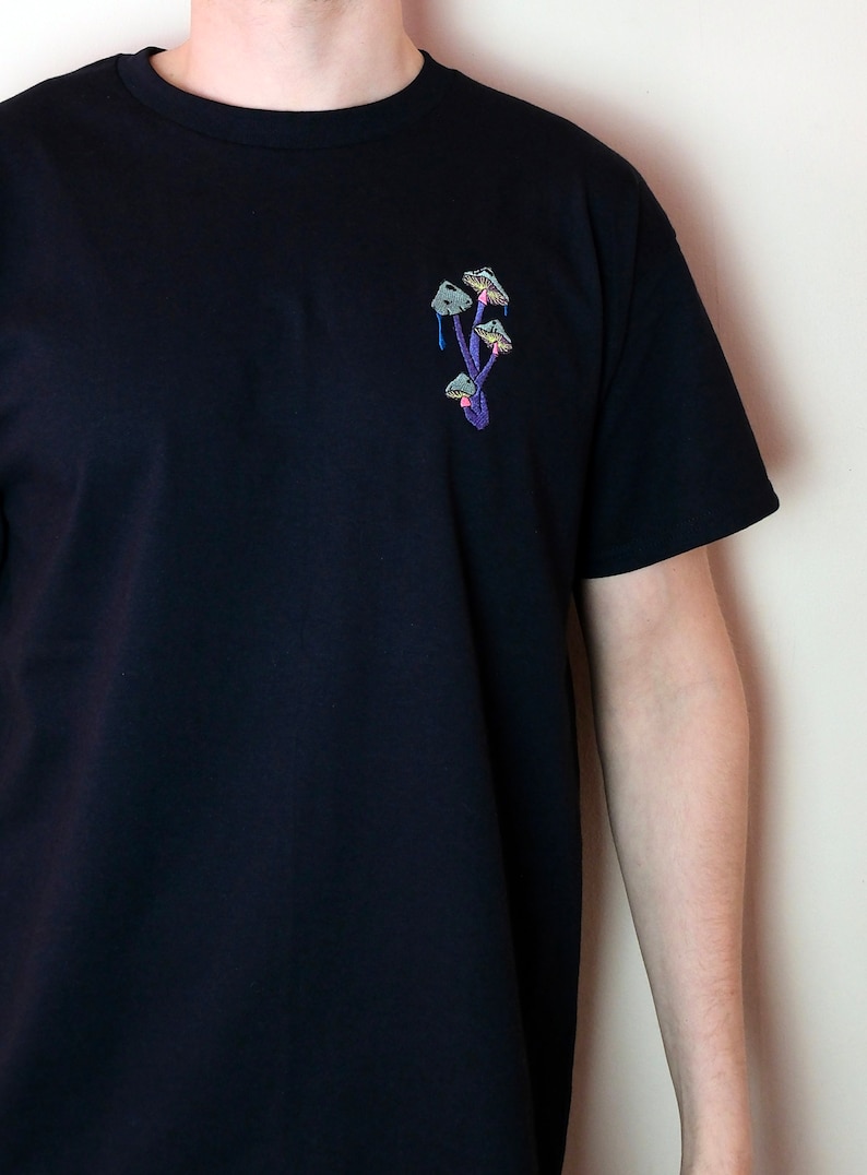 Peut inclure: Un t-shirt noir avec un motif brod&eacute; de champignons violets sur la poitrine gauche.