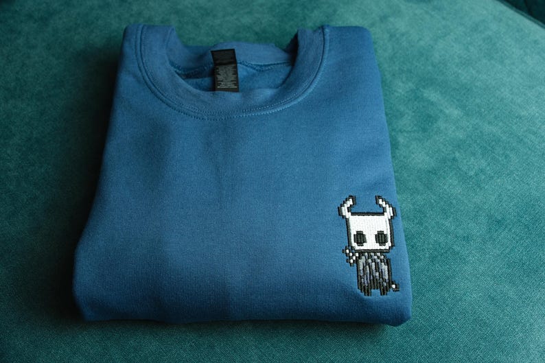 May include: A folded, teal-blue sweatshirt featuring a pixelated character design on the front. The design is a white and black character with a horned head. The sweatshirt is made of a soft material.