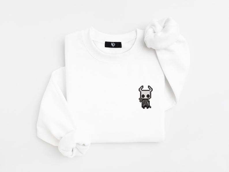 May include: White sweatshirt with a small, pixelated black and white character design on the front. The sweatshirt has long sleeves and a crew neck. A small black tag is visible near the neckline.