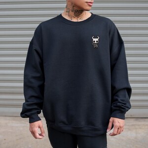 May include: Black crewneck sweatshirt with a small embroidered design on the chest. The design features a white and brown character. The sweatshirt is made of a soft material and has long sleeves.
