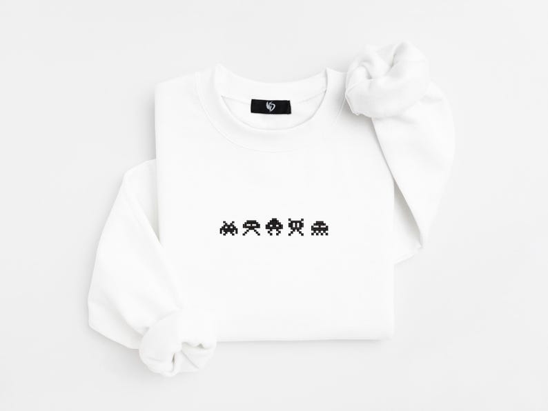 May include: White crew neck sweatshirt with a pixelated video game graphic across the chest. The graphic features four different alien-like characters in black. The sweatshirt has long sleeves and a small black tag near the neckline.