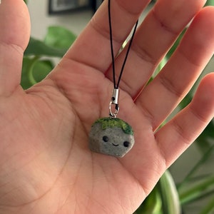 May include: A small gray and green stone charm with a happy face and a black string attached. The charm is being held in a person's hand.