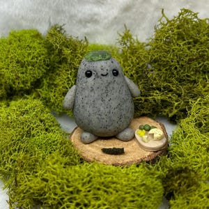 May include: A small, gray, speckled, rounded figurine with a smiling face and a green cap on its head. The figurine stands on a circular wooden slice next to a tiny, oval-shaped plate containing small, sculpted food items, including green and yellow pieces.