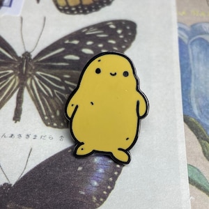 Könnte beinhalten: Ein gelber Emaille-Pin in Form einer lächelnden Kartoffel mit schwarzen Umrissen. Der Pin befindet sich vor einem Hintergrund aus Vintage-Schmetterlings- und Blumenillustrationen.
