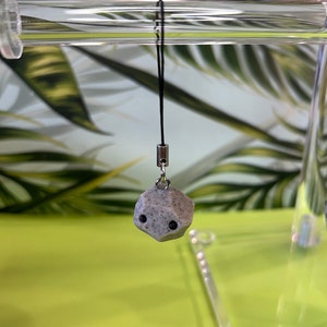 May include: A gray stone-like charm with black eyes hangs from a black string. The charm is attached to a silver ring and is hanging from a clear acrylic display stand.