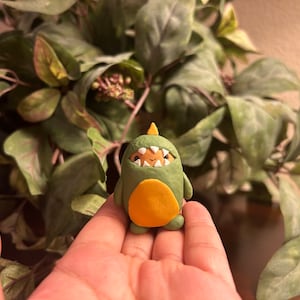 May include: A small, handmade green dinosaur figurine with an orange belly and a yellow horn. The dinosaur has a smiling face with visible teeth. The figurine is held in a person's hand, with a backdrop of green leaves.
