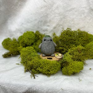 May include: A small, gray, speckled, rounded clay figurine with a smooth texture, two black dot eyes, and a tiny smile. The figurine has a green patch on its head and sits on a circular wooden base next to a miniature plate of food.