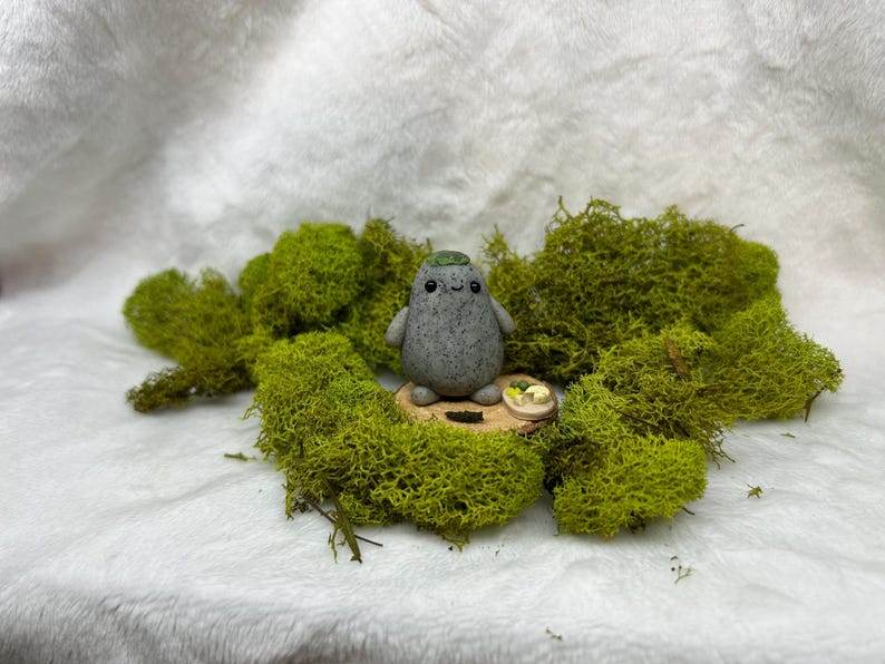 May include: A small, gray, speckled, rounded clay figurine with a smooth texture, two black dot eyes, and a tiny smile. The figurine has a green patch on its head and sits on a circular wooden base next to a miniature plate of food.