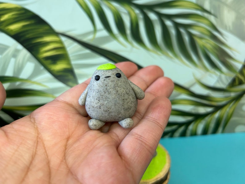 May include: A small, rounded figurine with a speckled grey, stone-like texture. The figure features two tiny arms, two small feet, two black dot eyes, a simple horizontal line for a mouth, and a smooth, bright lime green cap on its head.