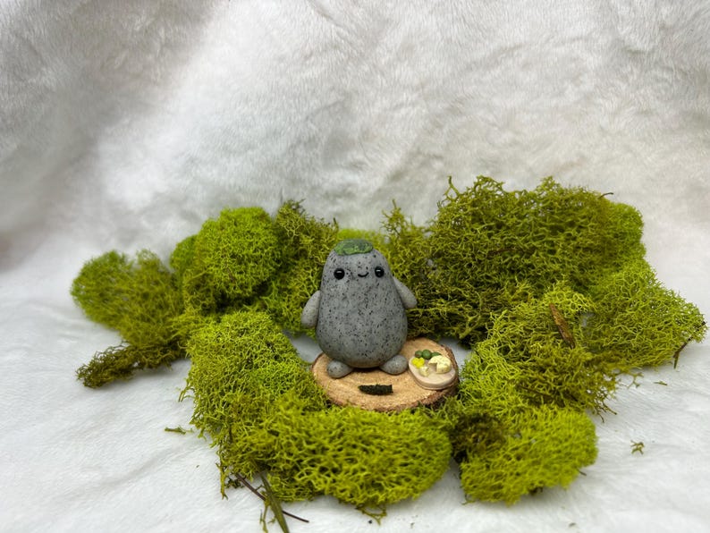 May include: A small, gray, speckled, rounded figurine with a smiling face and a green cap on its head. The figurine stands on a circular wooden slice next to a tiny, oval-shaped plate containing small, sculpted food items, including green and yellow pieces.