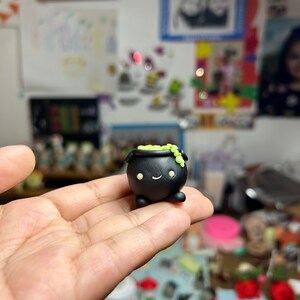 Peut inclure: Une figurine de petit chaudron noir avec un visage souriant et des d&eacute;tails verts. Le petit chaudron a un corps arrondi, de petites jambes et un bord rempli de d&eacute;corations vertes en forme de perles. Il est tenu dans la main d'une personne.