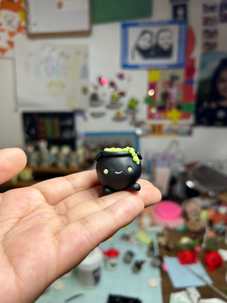 Peut inclure: Une figurine de petit chaudron noir avec un visage souriant et des d&eacute;tails verts. Le petit chaudron a un corps arrondi, de petites jambes et un bord rempli de d&eacute;corations vertes en forme de perles. Il est tenu dans la main d'une personne.