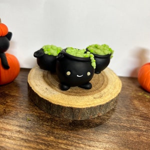 Peut inclure: Trois figurines de chaudrons noirs avec des visages souriants et un contenu vert bouillonnant sont pos&eacute;es sur une tranche de bois. Les chaudrons font partie d'une exposition sur le th&egrave;me d'Halloween, avec une citrouille et une figurine de chat noir visibles.