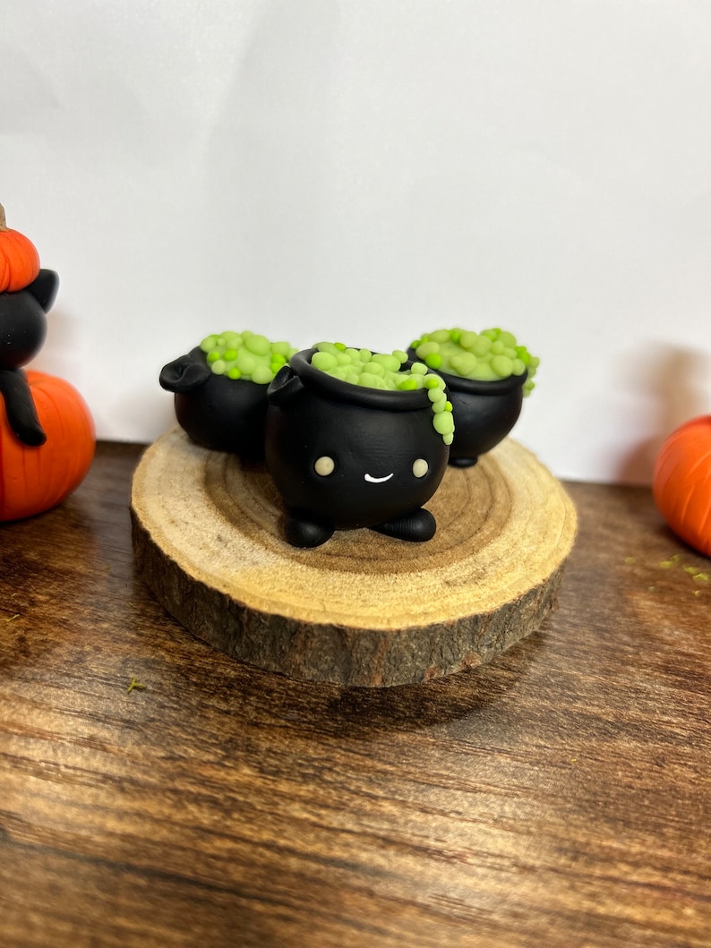 Peut inclure: Trois figurines de chaudrons noirs avec des visages souriants et un contenu vert bouillonnant sont pos&eacute;es sur une tranche de bois. Les chaudrons font partie d'une exposition sur le th&egrave;me d'Halloween, avec une citrouille et une figurine de chat noir visibles.