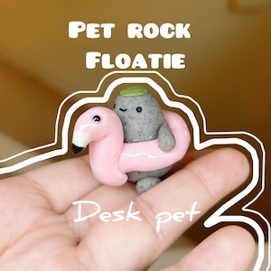 Puede incluir: Una pequeña roca gris con una hoja verde en la parte superior flota en un flotador de piscina rosa en forma de flamenco. La roca tiene una cara sonriente. El texto "Pet Rock Floatie" está por encima de la roca y el texto "Desk Pet" está debajo de la roca.