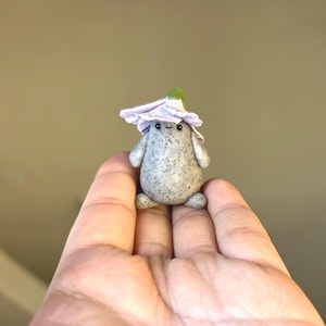 May include: A small, grey, stone-like figurine with a friendly face and a purple flower hat. The figurine is sitting on a hand.