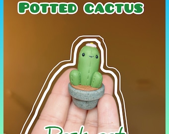 May include: A small green potted cactus with a white flower on top and a smiling face. The cactus is in a grey pot with a brown base. The text "Desk pet" is written below the cactus.