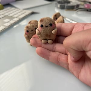 May include: A collection of small, brown, clay figurines shaped like smiling characters with rosy cheeks. Each figure has small arms and legs. The figurines are displayed on a light-colored surface.