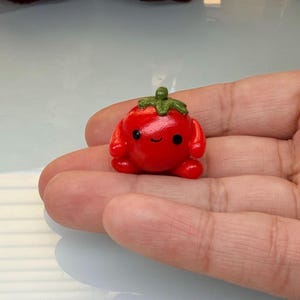 May include: A small, red, tomato-shaped figurine with a smiling face and green leafy top. The figurine is a cute, miniature sculpture, likely handmade, and is resting in the palm of a hand.