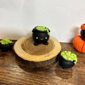 Peut inclure: Plusieurs d&eacute;corations miniatures sur le th&egrave;me d'Halloween. L'image pr&eacute;sente de petits chaudrons noirs remplis d'une potion verte et p&eacute;tillante. Un chaudron est plac&eacute; sur une tranche de bois, avec d'autres autour. Une citrouille orange est &eacute;galement visible.