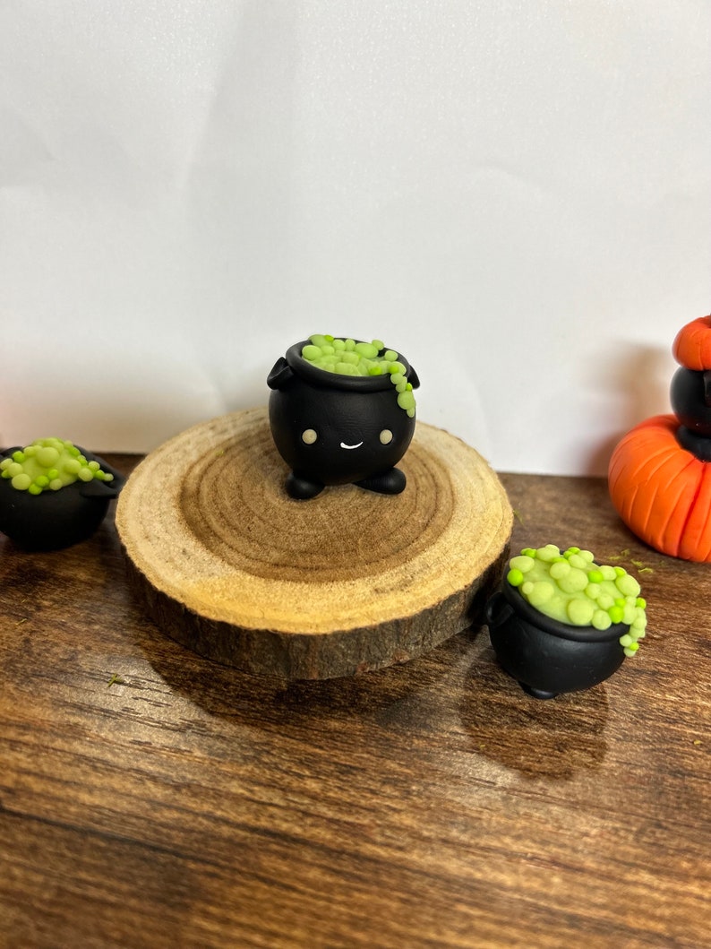 Peut inclure: Plusieurs d&eacute;corations miniatures sur le th&egrave;me d'Halloween. L'image pr&eacute;sente de petits chaudrons noirs remplis d'une potion verte et p&eacute;tillante. Un chaudron est plac&eacute; sur une tranche de bois, avec d'autres autour. Une citrouille orange est &eacute;galement visible.