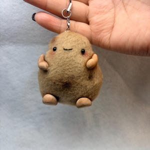 May include: A brown, fuzzy, round needle-felted potato charm with a black string loop. The potato features two small black bead eyes, a stitched smile, pink blush marks on the cheeks, and four small, smooth, light brown limbs attached to the body.