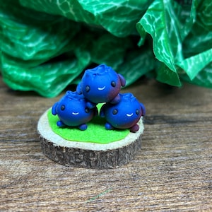 Puede incluir: Tres pequeñas figuritas de arcilla azul y púrpura con caras sonrientes, apiladas una encima de la otra sobre una base de madera verde y marrón.