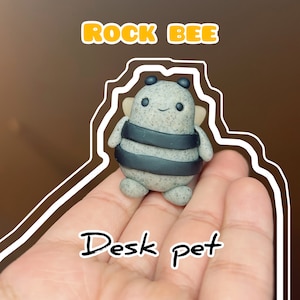 May include: A small, gray and black bee-shaped figurine with a friendly face. The figurine is made of a textured material and has a black and white striped body. The figurine is being held in a hand, and the text "Rock Bee Desk Pet" is written below it.